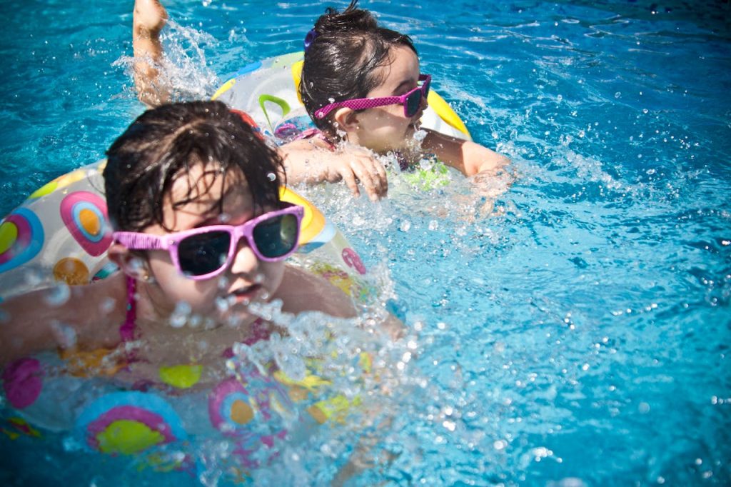 2-girl-s-swimming-during-daytime-61129 Two young girls enjoying a playful day in a bright blue swimming pool with colorful float rings.